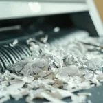 Trituradora de papel con papel atascado en las cuchillas, mostrando trozos de papel mal triturados y arrugados atorados en la entrada de la máquina. La imagen ilustra la importancia de mantener las cuchillas afiladas para evitar atascos y mejorar el rendimiento. La trituradora está sobre un escritorio, con hojas de papel intactas y herramientas como tijeras cerca, sugiriendo un intento de resolver el problema. Relacionado con métodos para afilar cuchillas y mantener la trituradora en óptimas condiciones.